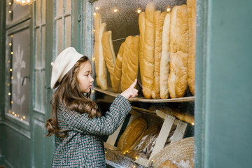 A stylish, beautiful six-year-old girl in a white beret and green checked coat points to a baguette in a bakery window