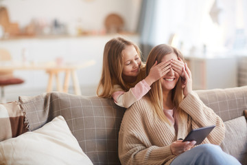 Warm toned portrait of cute girl playing peek a boo with happy young mother while surprising her on Mothers day, copy space