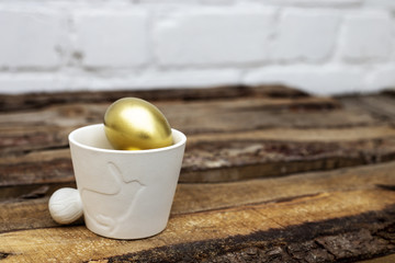 White pot with decorative bunny and Easter golden eggs on brown wooden table and white bricked background. Easter rabbit, eco decoration, spring. Copy space
