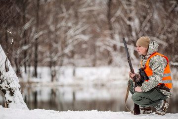 Male hunter in camouflage, armed with a rifle, standing in a snowy winter forest with duck prey