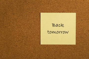 Small yellow sticky note on an office cork bulletin board with the message back tomorrow