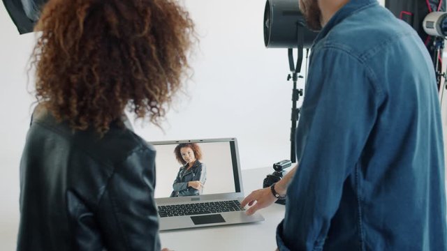Photographer bearded guy is showing photos to mixed race model using laptop computer in studio talking pointing at screen. Business and devices concept.