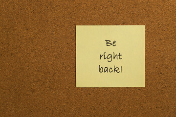Small yellow sticky note on an office cork bulletin board with the message be right back