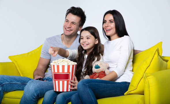 Favorite moment. Beautiful parents and their cute little daughter are laughing and eating popcorn, while the kid is pointing at TV.