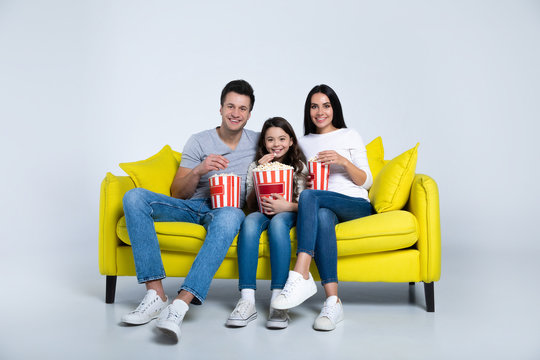 Movie Night Snack. A Small Cheerful Family Sitting On A Yellow Sofa In Casual Clothes, Eating Popcorn And Smiling, While Looking In The Camera.