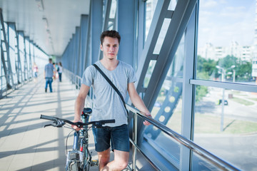 Front view of young man with bicycle holding handrail on bridge and looking at camera. Good-looking guy with bike wearing t-shirt and jean shorts. Concept of bicycling and man in city.