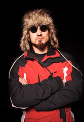 Front view of handsome gentleman with crossed arms looking at camera. Good-looking guy in sunglasses wearing fur hat and jacket. Isolated on black studio background. Concept of men fashion.