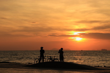 The silhouette of lovers riding bicycles and watching and photographing the sunrise on the beach at dawn