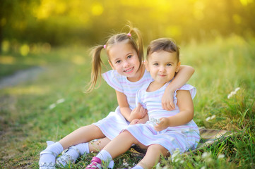 Cute children are walking in the park at sunset. Girls gently hug while sitting on the grass. Children play and have fun outdoors.
