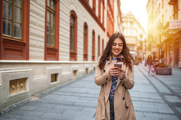 Fototapeta premium Girl walking and texting on the smart phone in the street. Happy Latin woman smiling brightly while holding smart phone strolling outdoors in the city at sunny day,female person using mobile