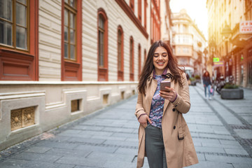 Fototapeta premium Gorgeous beautiful young woman with brown hair messaging on the smart-phone at the city street background. pretty girl having smart phone conversation in sun flare.