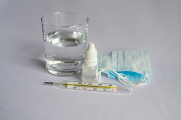 A glass of water, a thermometer with a high temperature indicator, a medical mask, pills, drops for a runny nose. White background, no one there