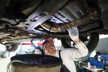 Mechanic conducts thorough inspection car garage. Male mechanic in working overall stands under car and shines lantern on car parts in search malfunction. Technician smiles while draining old oil. © H_Ko
