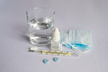 A glass of water, a thermometer with a high temperature indicator, a medical mask, pills, drops for a runny nose. White background, no one there