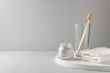 Fototapeta na wymiar Bamboo toothbrush in a glass cup, next to whitea cotton towel and powder for brushing your teeth in jar. Light wooden surface, gray backdrop. Biodegradable personal care products. No plastic concept.