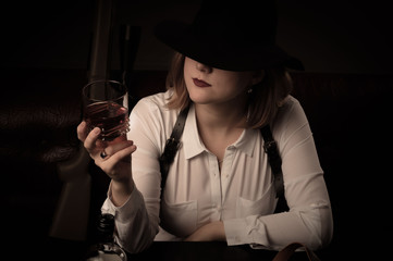Retro mafia woman in a white blouse and a black hat sits at a table with air guns. Whiskey in a glass. - obrazy, fototapety, plakaty