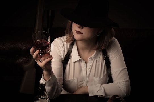 Retro Mafia Woman In A White Blouse And A Black Hat Sits At A Table With Air Guns. Whiskey In A Glass.