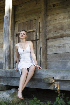 A girl in a white dress is sitting barefoot, on the wooden threshold of an old farmhouse