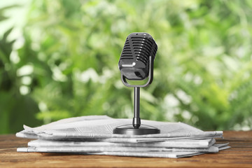 Newspapers and vintage microphone on wooden table against blurred green background. Journalist's work