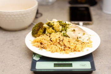 Bulgur, white fish and vegetables on a kitchen scale. Delicious healthy warm salad on a marble table. Bulgur pilaf. Counting calories. Close up.