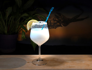 white cocktail with lemon and straw on dark backdrop with orange glow and leaves. Edge of tea glass with sugar.