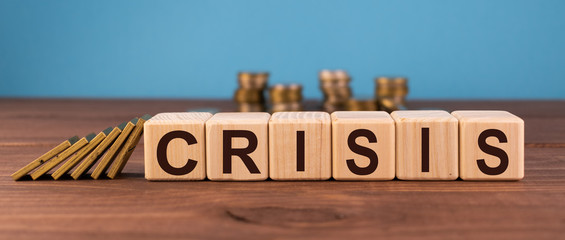 Word Crisis on wooden cube blocks on wood table with stacks of coins on blue background. Financial crisis, investment or economic bubble burst countdown concept.