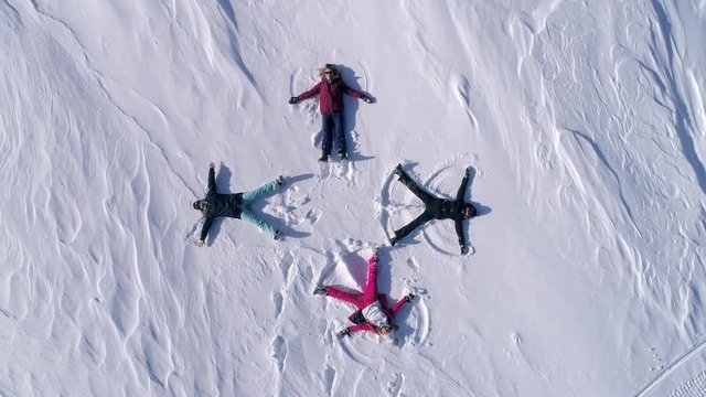 AERIAL TOP UP: a group of young people in bright ski suits depict snow angels lying on the fresh snow in the form of a cross. four winter angels happy and energetic with splashes of snow. SLOW MOTION