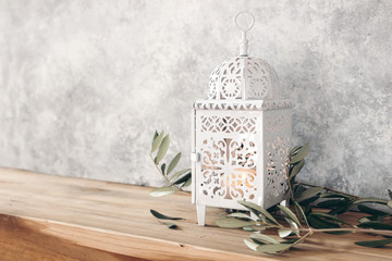 White ornamental Moroccan, Arabic lantern. Green olive leaves, branches on old wooden table, blurred grunge wall background,. Greeting card for Muslim holiday Ramadan Kareem. Festive still life.