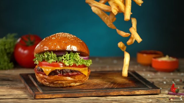 Super Slow Motion Shot of Fast Food Concept. Falling French Fries on Wooden Cutting Board next to the Fresh Hamburger at 1000fps.