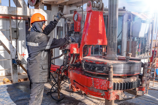 Work driller in red uniform, in helmet and goggles. He uses a hydraulic wrench to screw drill pipes to lower them into an oil well and continue drilling it. The concept of a working person.