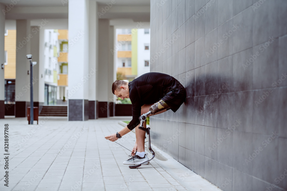 Wall mural Side view of attractive caucasian sportsman with artificial leg leaning on the wall and tying his shoelace. - Wall murals