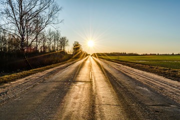 The asphalt of an old road, cracked in the cold, is lit by the sun's rays in a haze of blue sky against the background of a overed fields and trees