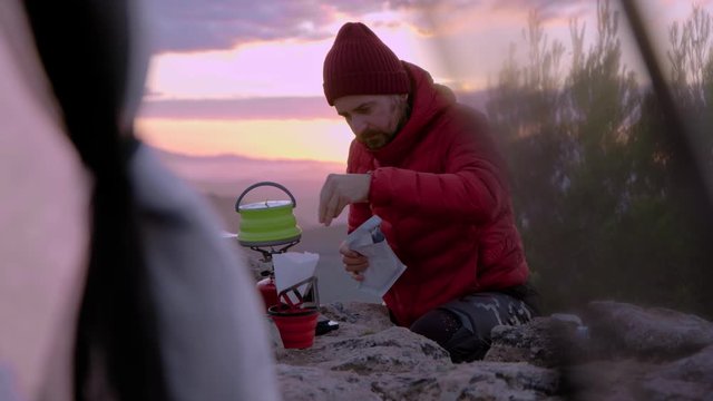 Beautiful cinematic shot in pink and purple tones of man prepare coffee and breakfast outdoors on gas camping stove at sunrise or sunset. Tourist cooking outside near tent