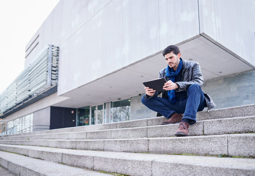 young man sitting on the outside stairs of a public library using his tablet computer