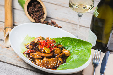 Sliced chicken fried in sweet and sour soy sauce with vegetables and herbs in a deep plate on a wooden table in a restaurant.