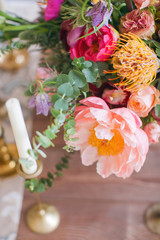 A bouquet of blooming peony, roses and other flowers in a vase on a rustic table surrounded by candles with vintage candlesticks. Boho chic for the holiday. Scenery decoration for dinner.