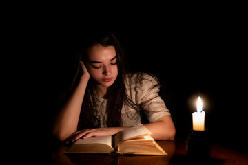 A young girl sleeping over an old book at night with candle light. The girl is wearing an old white dress. Dark background. Front view.