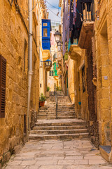 Old narrow medieval street with yellow buildings with colorful balconies in town Singlea, Valletta, Malta - obrazy, fototapety, plakaty