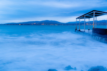 Storm waves at high speed run over the stones of Gelendzhik beach. Waves due to exposure are a blue fog. The resort of Gelendzhik, the evening twilight. 