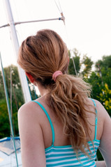 back view of beautiful woman in blue top with pony tail looking away in front of sky and green trees in yachts club