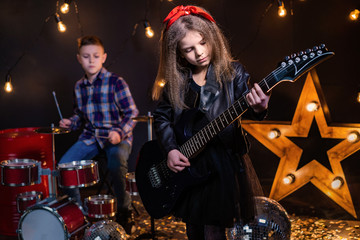 Kids pretending to be in a rock band and play and sing at studio or stage. Girl play on guitar and boy on drums.