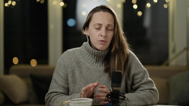 Young woman in a gray sweater records at home a podcast on a background of lights