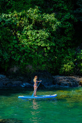 Fototapeta premium SUP Stand up paddle board. Blond girl sailing on paddle board in the green sea near stone beach. Woman on summer holidays vacation lifestyle. Mtsvane Kontskhi Beach, Batumi, Georgia.
