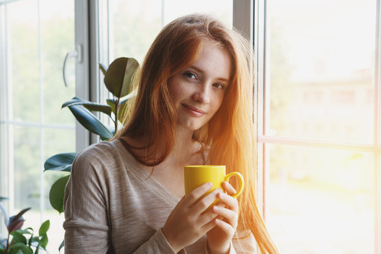 candid portrait of beautiful cheerful young smiling red hair woman holding yellow coffee cup, enjoying morning coffee at home
