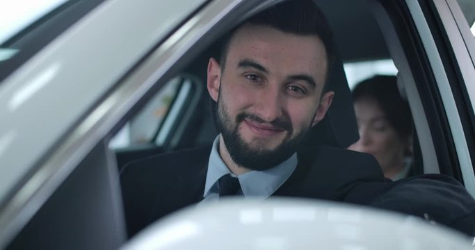 Portrait of positive young Caucasian man sitting in car with passenger and looking at camera. Smiling professional chauffeur working in taxi or as private driver. Cinema 4k ProRes HQ.