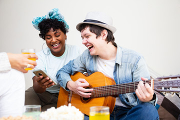 Friends in their twenties playing guitar and singing together in living room with food snacks and drinks. Playful youth, enjoyment situation .