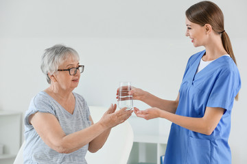 Female doctor giving senior woman suffering from Parkinson syndrome glass of water in clinic