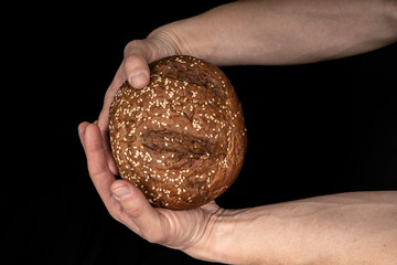 Man's hands hold tasty fresh loaf of dark bread with sesame seeds on black background