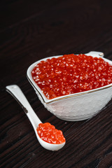Red caviar in a white ceramic bowl with spoon on a brown rustic wooden background