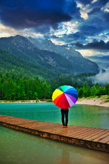 Unrecognizable person with colorful rainbow umbrella standing in the rain on a wooden pier on a...
